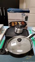 Photo showing Rival Gourmet Fold n Store griddle with ridged surface, Bravetti slow heater placed on top, and black skillet with glass lid in front.