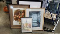 Four framed artworks stacked together showing variety in style and subject matter including floral, village street, hallway interior.