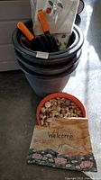Photo shows the stacked 4 black plastic planters, small terracotta pot with polished rocks, garden tools, and white cloth.