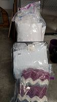 Photo showing four pillows in plastic bags stacked behind a double size quilt in sealed plastic with a hand-crocheted afghan in front.