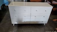 Front and top view of white wooden dresser with 6 drawers, resting on short tapered legs.