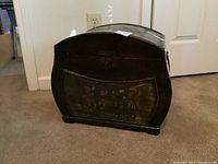 Front view of curved wood chest with ornate embossed metal panel showing cherubs and floral design, and hasp closure on top.
