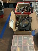 Box with assorted hand tools including multiple screwdrivers, pliers, wrenches, and a large tabletop magnifying glass