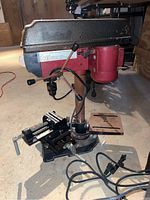 Front-left angled view of the red and black 5-speed mini drill press on a workshop floor showing motor housing, safety guard, and adjustable vice on base.