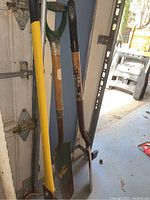 Photo of several garden tools including shovels and post hole digger with wooden and yellow handles leaning upright