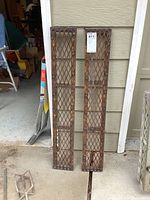 Front view of two rectangular metal grate ramps leaning side by side against a wall outdoors. Both ramps show rust and weathering.