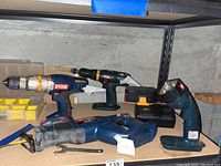 Photo showing three Ryobi power tools including a cordless drill driver, reciprocating saw, flashlight on a shelf with battery charger and battery.