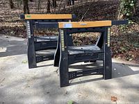 Two black plastic adjustable sawhorses placed outdoors on a concrete surface with wooded background. Both sawhorses have yellow strips on sides showing adjustment angles from 30 to 37 inches.