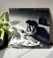 Front view of Billy Joel The Stranger album cover in natural light showing Billy Joel holding a mask on a bed.