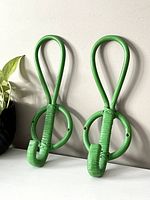 Two green wicker wall hooks arranged vertically, resting against a wall, showing their size and looped top design with circular base detail.