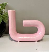 Front view of a pink ceramic vase with a curved right-angle tubular shape on white surface with a plant in the background.