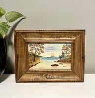 Front view of the small framed hand-painted sailboat picture sitting on a white surface with a green plant nearby.