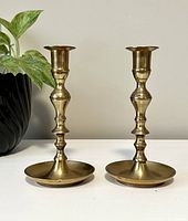 Front view of both vintage brass candle stick holders placed side by side on a white surface with a green plant in background.