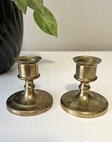 Front view of two small vintage brass candle stick holders with patina and visible wear from age, placed side by side on a white surface with a plant in the background.