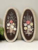 Pair of oval vintage chalkware floral wall hangings with carved cream frames and dark brown textured backdrops displaying raised flower bouquets