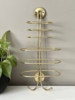 Front view of vintage brass toned metal towel rack showing five horizontal towel bars and two hooks at the bottom against a plain wall, alongside a green potted plant.
