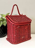 Front angled view of red wicker letterbox showing curved handle, closed lid and woven texture with some paint wear.