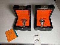 Two Burett Swiss watches on orange pillows inside open black wooden boxes with Burett branding, accompanied by an orange booklet and lot number card.