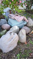Photo showing 11 hollow fake rocks in different shapes and colors along with 3 folded shade cloths in green, pink, and blue/grey colors placed on grass near shrubs.