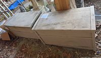 Two weathered beige Suncast outdoor storage deck boxes side by side on dirt ground under a covered area, tops visibly stained and worn.