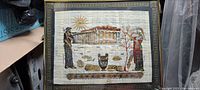 Framed papyrus artwork showing two robed figures flanking a large ancient Greek temple, with a vase and rocks in the foreground and a decorative border.