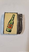 Top view showing 7 Up bottle illustration on cream-colored panel with metal lighter mechanism attached.