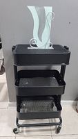 Front view of black metal RÅSKOG utility cart with three mesh baskets and decorative item on top.