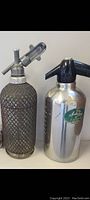 Photo of two vintage Sparklets makers seltzer bottles side by side, one with a textured glass body and metal dispenser, the other metallic with black plastic spout and green label.