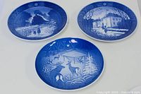 Three vintage Royal Copenhagen Christmas plates arranged on white background showing winter scenes with people and reindeer