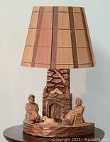 Full front view of carved wood lamp showing detailed base with two seated figures and stone fireplace, topped with wood panel lampshade.