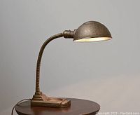 Side view of the lamp on a round wooden table showing the dome-shaped shade, flexible gooseneck, and cast iron base.