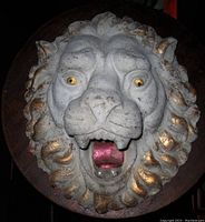 Close-up frontal image of the cast stone lion head wall sculpture, showing details of the face, mane, painted eyes, and open mouth with teeth and tongue.