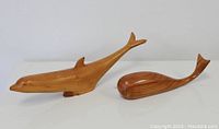 Two hand-carved wooden marine animal sculptures, a dolphin and a whale, shown from a side perspective on a white surface.