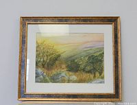 Framed watercolor painting depicting a scenic autumn/yellow field landscape with trees and distant hills, mounted on a wall.
