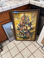 Framed abstract tree painting leaning against kitchen cabinets on tiled floor alongside three other framed artworks.