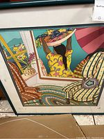 Close-up photo of framed artwork showing woman carrying basket on head and colorful furniture indoors.