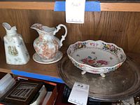 Three porcelain items including two pitchers and one footed bowl displayed on wooden shelf with price tag.