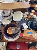 Top view of assorted kitchenware including large ceramic bowls with fruit designs, white bowls, plastic containers, ceramic mugs, and utensils in wooden holder. Includes a metal strainer on a set of colorful bowls.