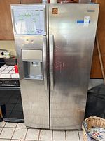 Front view of the stainless steel Frigidaire side-by-side refrigerator with water and ice dispenser on left door. Visible dents, scratches, and attached whiteboard on the left door.