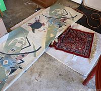 Three rug runners lying rolled and flat on a floor showing designs and condition; one runner features abstract geometric and tribal motifs, another is a smaller, red traditional style rug with floral patterns and fringed edges; the third is a light-colored runner.