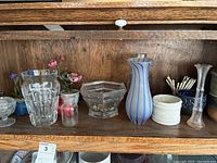 Shelf with 8 items including clear faceted glass vase, hexagonal glass bowl, blue striped vase, small ceramic pot with artificial flowers, white ceramic bowl, small glass container with frosted lid and black top, small clear glass vase with flared rim, and small blue bowl with decorative sticks.