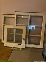 Set of three antique wood frame windows with glass panes shown stacked against a wall, displaying different sizes and visible wear on painted surfaces.
