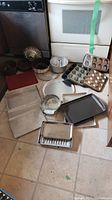 Wide view showing a variety of baking items including muffin tins, Bundt pans, pizza pan, grill pans, cookie sheets, and baking trays lined on floor.
