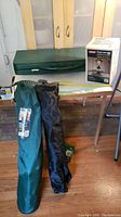 Photo showing folded green camping table on wooden floor with green and black carrying bags for chairs and boxed propane lantern on top of table.