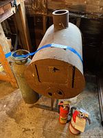 Front angle view of vintage cylindrical cast iron portable stove on metal stand with chimney pipe and three fire extinguishers placed below it, two small red and white extinguishers and one tall antique extinguisher.