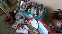 Overall display of assorted Christmas decorations showing snowman cookie jar, Santa plate, ornaments, and village figurines on a floor and a green plastic container.
