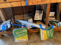 Painting supplies displayed on a wooden shelf, showing tarps in packaging, plastic drop sheet roll, and various tools underneath shelf