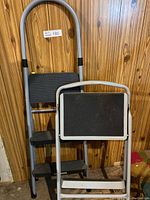 Two gray metal step ladders with black steps, one with three steps, the other partially visible with two steps, against wood panel wall.