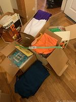 Photo showing multiple boxes with folded assorted fabrics in solid colors such as purple, white, orange, teal, black, cream, and gray placed on a wooden floor.