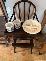 Antique wooden child's highchair with spindle backrest and footrest, two children's cups with colorful illustrations, and a vintage divided children's plate with red designs are arranged together.